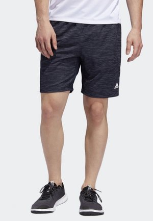 Mand iført sorte atletikshorts med diskret mønster og hvid Adidas-logo, kombineret med sorte løbesko og hvid skjorte.
