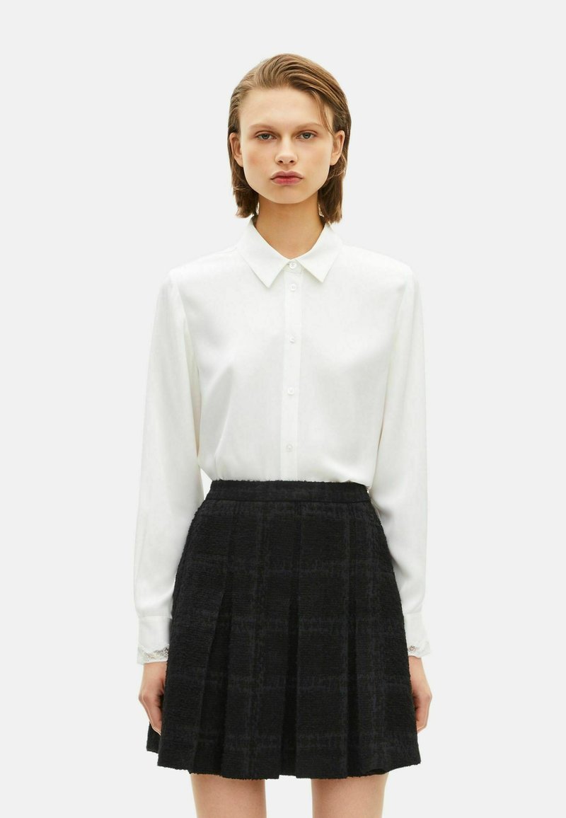 Witte blouse met lange mouwen, een puntkraag en knoopsluiting, gecombineerd met een zwarte plissérok met een gestructureerd patroon.