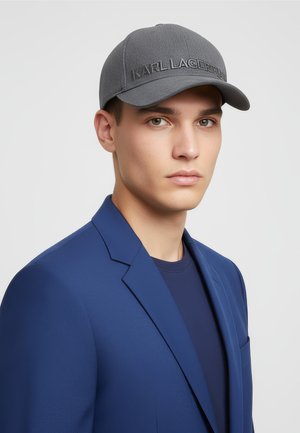 Jeune homme portant une casquette grise Karl Lagerfeld, un blazer bleu et une chemise bleu foncé, faisant face à l'avant avec une expression neutre sur un fond blanc.