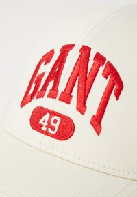 Căciulă de baseball crem cu text brodat îndrăzneț roșu "GANT 49", având un design structurat, vizieră curbată și material texturat.