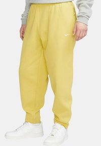 Gula, löst sittande träningsoverallsbyxor med elastisk midja, sidofickor och Nike-logotyp. Bärs med vita sneakers. Mjukt tyg i texturen.