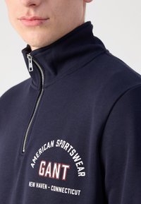 Navy blue zip-up sweater made of smooth fabric, featuring a high collar, white text logo "GANT," and a close-fitting design.