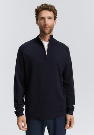 Man met krullend haar in een donkerblauwe rits-vrij pullover over een wit shirt, staande tegen een effen lichte achtergrond.