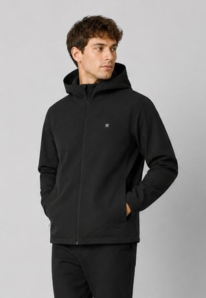 Homme portant un sweat à capuche noir zippé, les mains dans les poches, regardant sur le côté sur un fond gris clair uni.