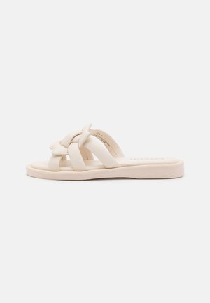 Beige flad slidesandal med polstrede flettede remme og en blød polstret sål, vist fra siden på en ensfarvet baggrund.