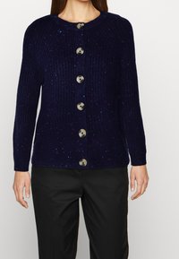 Marinblå ribbad cardigan med flerfärgade prickar, med sex stora runda knappar fram och långa ärmar. Slät stickad textur.