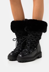 Bottes d'hiver noires mi-mollet avec lacets à l'avant, tige en tissu à motif, semelle épaisse en caoutchouc, et bordure en fausse fourrure retroussée portées sur jambes nues.