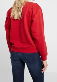 Sudadera roja de corte relajado, con puños y cinturilla de canalé. Combinada con jeans ajustados de color azul oscuro. Vista desde la parte de atrás.