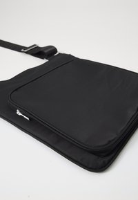 Borsa a tracolla nera con una texture in tessuto liscia, dotata di una tasca frontale con zip e una tracolla regolabile. Design rettangolare semplice.