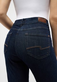Mørkeblå dongeribukser med en tettsittende design, med baklommer med buet stitching og en brun lærmerke.