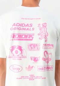 Hvit bomull t-skjorte med forskjellige rosa grafiske trykk, inkludert logoer og tekst knyttet til Adidas, navn på byer og designelementer.
