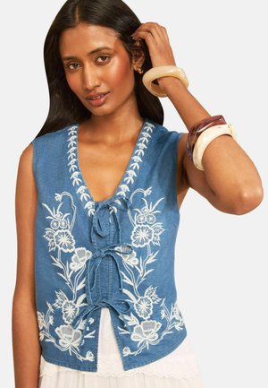 Kvinde iført blå ærmeløs top med hvid blomsterbroderi og tre bindebånd foran, accessoriseret med store armbånd på hendes hævede arm.