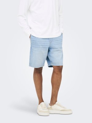 Homme portant un short en jean bleu clair, une chemise blanche à manches longues avec les mains dans les poches, et des baskets blanches, debout devant un fond uni.