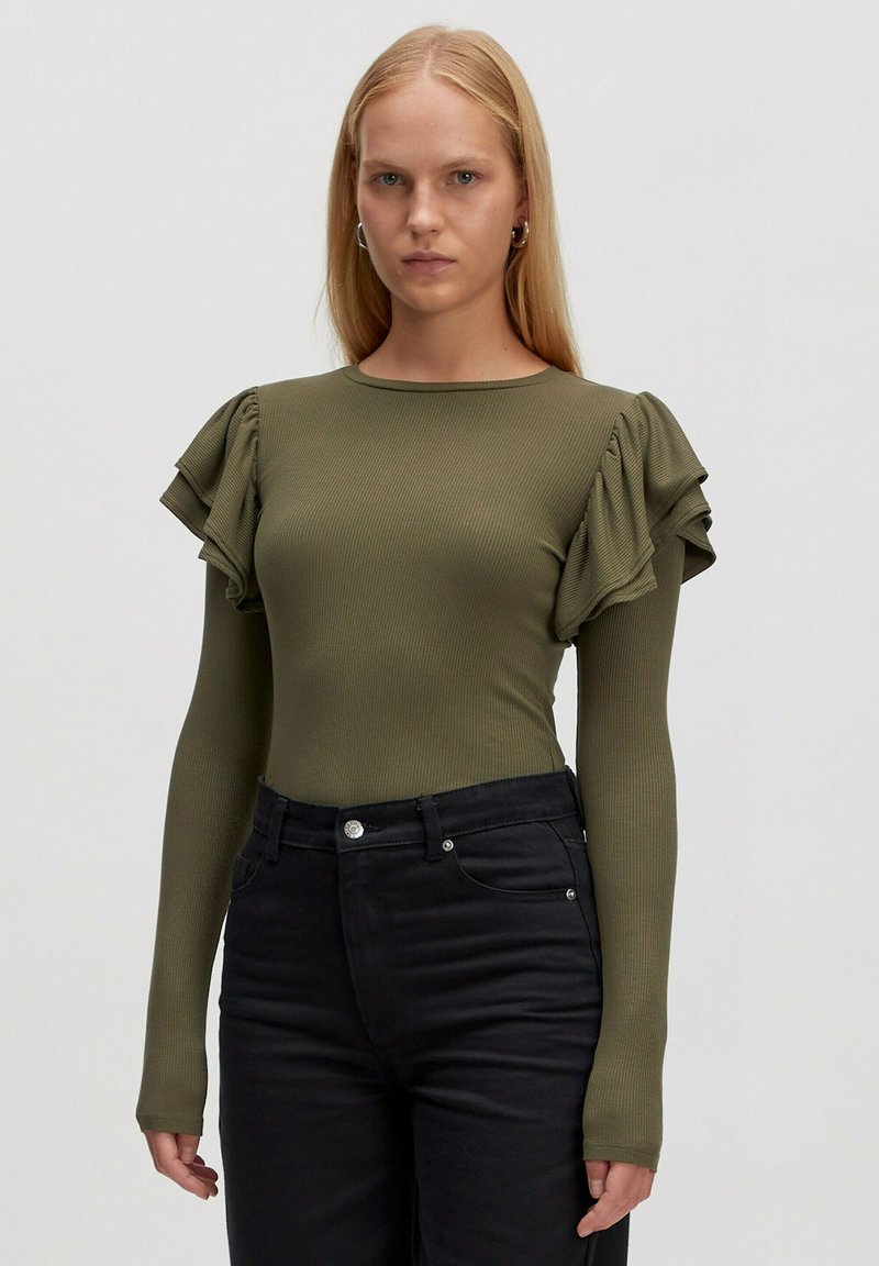 Olijfgroen geribbeld langemouwtop met ruches aan de korte mouwen, gecombineerd met zwarte aangesloten jeans en een zilveren knoopdetail op de taille.