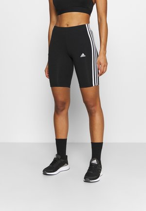 Kobieta w czarnych dopasowanych krótkich spodenkach Adidas z białymi bocznymi paskami, czarnym sportowym biustonoszu, czarnych skarpetkach i czarnych butach sportowych Adidas, stojąca na szarej podłodze.