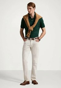 Mężczyzna w zielonej koszulce polo, białych spodniach, brązowym pasku, brązowych skórzanych butach i beżowym swetrze zarzuconym na ramionach, na jednolitym tle.