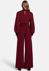 Burgundijas jumpsuit ar augstu kaklu, garām puff piedurknēm, akcentētu vidukli un plašu kāju siluetu. Aizmugurē ir izgriezums taustiņam.