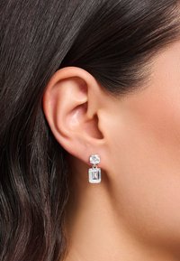 Silberne Drop-Ohrringe mit einem runden, klaren Edelstein oben und einem rechteckigen, klaren Edelstein darunter, die das Licht mit einer glatten Oberfläche reflektieren.