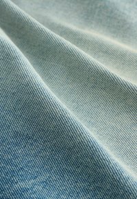 Heled sinine denimpindel, millel on sile tekstuur, nähtav kudumise mustrit ja peen kulumine, näidates volditud kihti ja pehmeid kontuure.