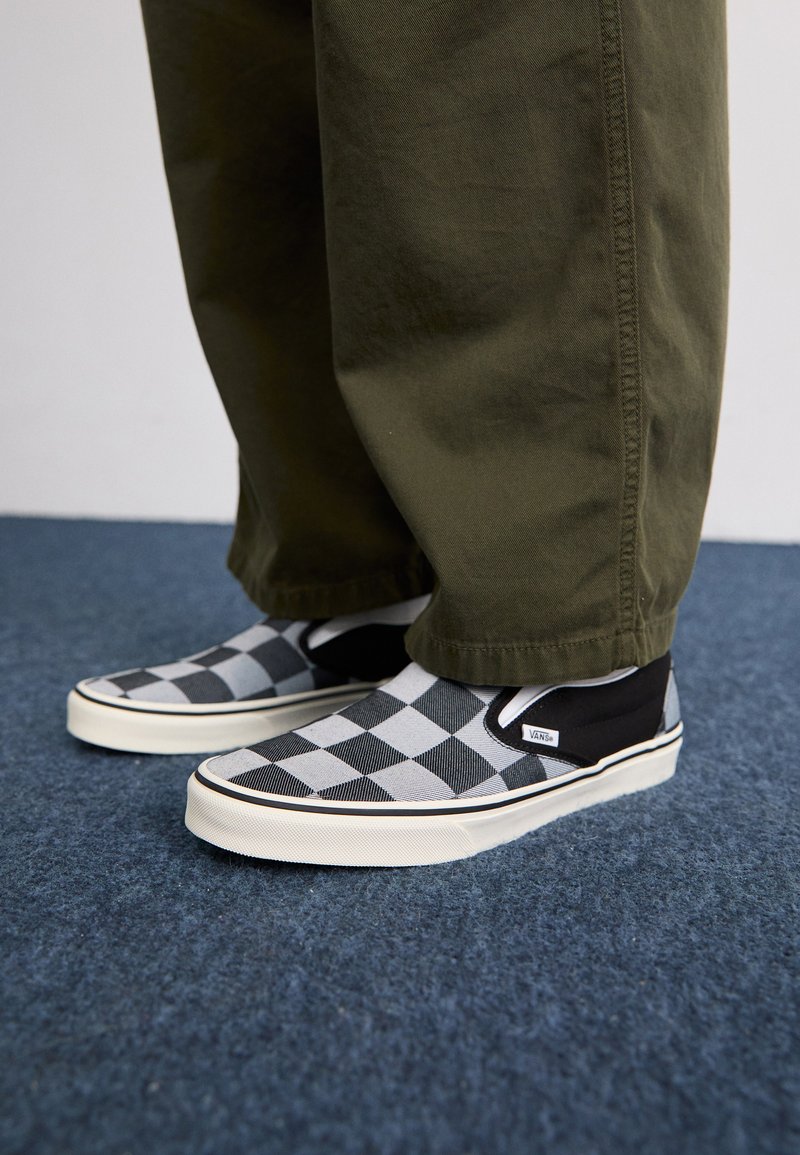 Zapatos slip-on en blanco y negro con un diseño de tablero de ajedrez, textura suave, acentos negros contrastantes y una suela blanca gruesa. Combinados con pantalones en color verde oliva.