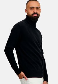 Pull en col roulé noir avec une texture douce, manches longues et poignets côtelés, porté avec un pantalon blanc. Des détails de couture subtils sont visibles.
