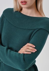Maglione teal a maglia con un ampio scollo a spalle scoperte e maniche lunghe. La texture è morbida e leggermente pelosa, con una forma aderente.