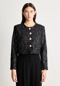 Giacca nera cropped con un motivo in pizzo floreale, dotata di sei bottoni rotondi, maniche lunghe e una finitura texturizzata. Abbinata a pantaloni neri.