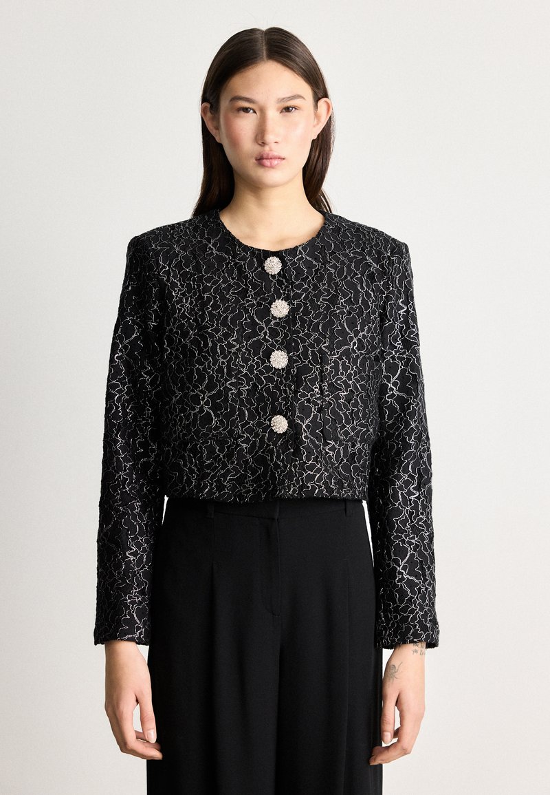Giacca nera cropped con un motivo in pizzo floreale, dotata di sei bottoni rotondi, maniche lunghe e una finitura texturizzata. Abbinata a pantaloni neri.