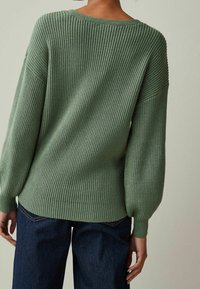 Pull vert en maille avec une texture côtelée, manches larges et coupe décontractée, associé à un jean bleu foncé. Détail de couture visible aux coutures.
