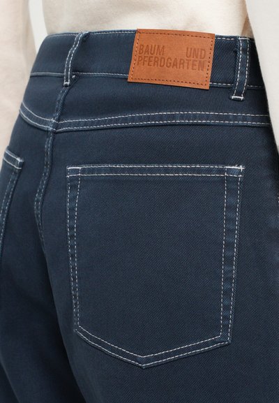 Tmavomodré denimové džínsy s svetlým prešitím, vybavené dvoma zadnými vreckami a obdĺžnikovou hnedou koženou nášivkou s označením "BAUM UND PFERDGARTEN."