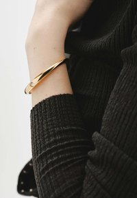 Pulsera de oro con acabado liso y un único acento de gema clara, llevada sobre la manga de un suéter negro texturizado.