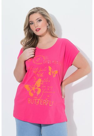 Kobieta o długich blond włosach, ubrana w jaskraworóżowy T-shirt z pomarańczowym motylem i napisem, w połączeniu z jasnoniebieskimi spodniami, stojąca przy białej ścianie.