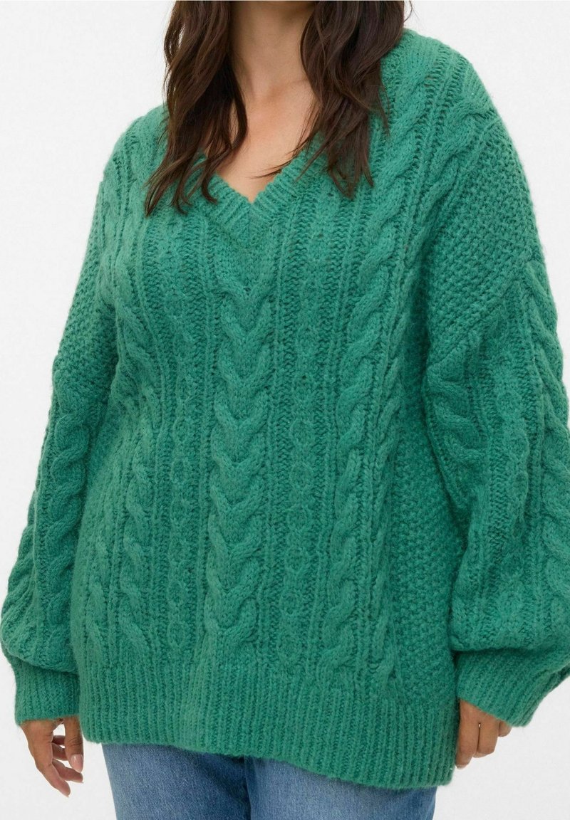 Femme portant un pull vert oversize en maille torsadée avec un col en V et des manches longues, assorti à un jean bleu.