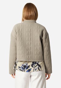 Gestrickte beige Jacke mit strukturierter Oberfläche, mit hohem Kragen und verkürzter Länge, die über ein floral bedrucktes Hemd getragen wird.