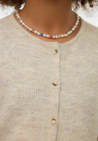 Béžový žebrovaný cardigan s kulatým výstřihem, zdobený světle hnědými knoflíky. Nošený s vícebarevným náhrdelníkem z perel a korálků.