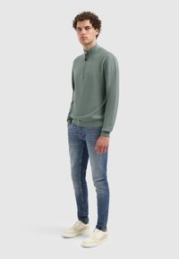 Groene gebreide pullover met een opstaande kraag en ritsdetails, gedragen met lichtblauwe jeans en crèmekleurige sneakers met grijze accenten.