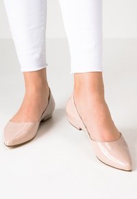 Nohy v nahých lakovaných balerínách s špičatou špičkou spojené s bílými zkrácenými skinny kalhotami na jednobarevném světlém pozadí.