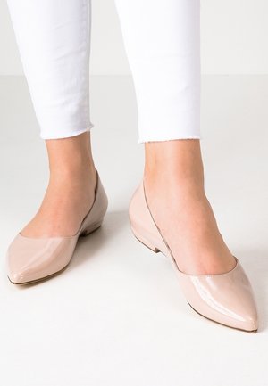 Piedi che indossano ballerine con punta affusolata in vernice color nude abbinate a pantaloni skinny bianchi a cervo su uno sfondo chiaro e semplice.