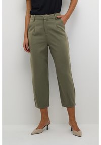 Pantalon ajusté vert olive à taille haute, longueur cropped et plis avant. Porté avec un haut noir et des talons beiges pointus.
