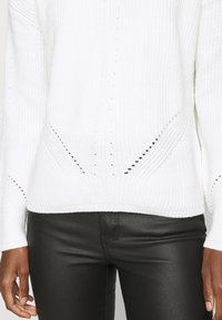 Persoon in een wit gebreid sweater met kleine geperforeerde patronen en zwarte slim-fit leren broek, met de handen langs het lichaam.