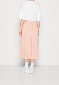 Vaaleanpunainen pleatsilta midi-hame, jossa on joustava vyötärö, yhdistetty valkoiseen lyhyt-hihaiseen t-paitaan ja valkoisiin lenkkareihin, nähtynä takaa.