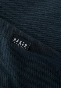 Tamsiai mėlynas tekstūruotas audinys su audimo raštu, kuriame matomas etiketas su užrašu "BAKER by Ted Baker" kontrastinga balta spalva.
