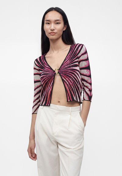 Femme portant un cardigan court coloré rose et noir à motifs, manches trois quarts, et un pantalon blanc taille haute, sur un fond uni.