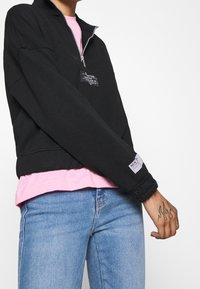 Svart zip-up sweatshirt med hög krage, med en framficka och elastiska ärmslut, lagd över en rosa skjorta och parat med ljusblå jeans.