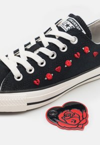 Μαύρο καμβά παπούτσι με λευκές κορδόνες, διακοσμημένο με κόκκινες κεντητές τριαντάφυλλα και καρδιές. Συνημμένο είναι μια γυαλιστερή κόκκινη καρδιά-σήμα με σχέδιο τριαντάφυλλου.