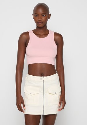 Kobieta z ogoloną głową, ubrana w jasnoróżowy bezrękawnik typu crop top oraz białą mini spódniczkę z przednim zamkiem błyskawicznym i dużymi kieszeniami.
