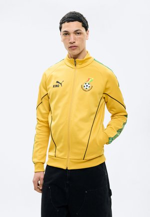 Tânăr purtând o jachetă galbenă a Asociației de Fotbal din Ghana cu pantaloni negri, stând cu o mână în buzunar pe un fundal alb.