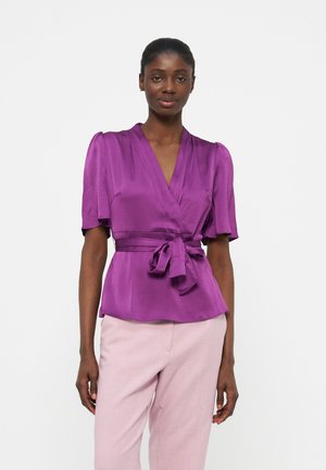 Femme portant un chemisier cache-coeur violet avec des manches courtes bouffantes et une taille nouée, assorti à un pantalon rose clair, se tenant devant un fond blanc.
