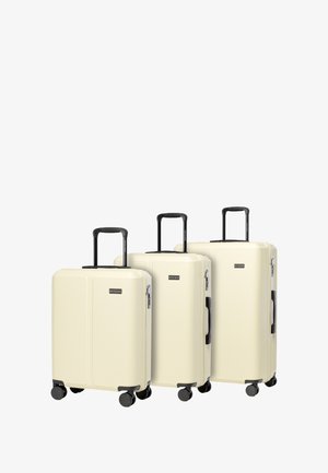 Drie harde koffers in crème, met een verticale geribbelde textuur, zwarte handvatten en vier draaibare wielen voor gemakkelijke mobiliteit.