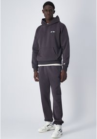Felpa in cotone grigio scuro con tasca frontale e un design grafico a occhio. Abbinata a pantaloni tuta grigio scuro e sneakers bianche.